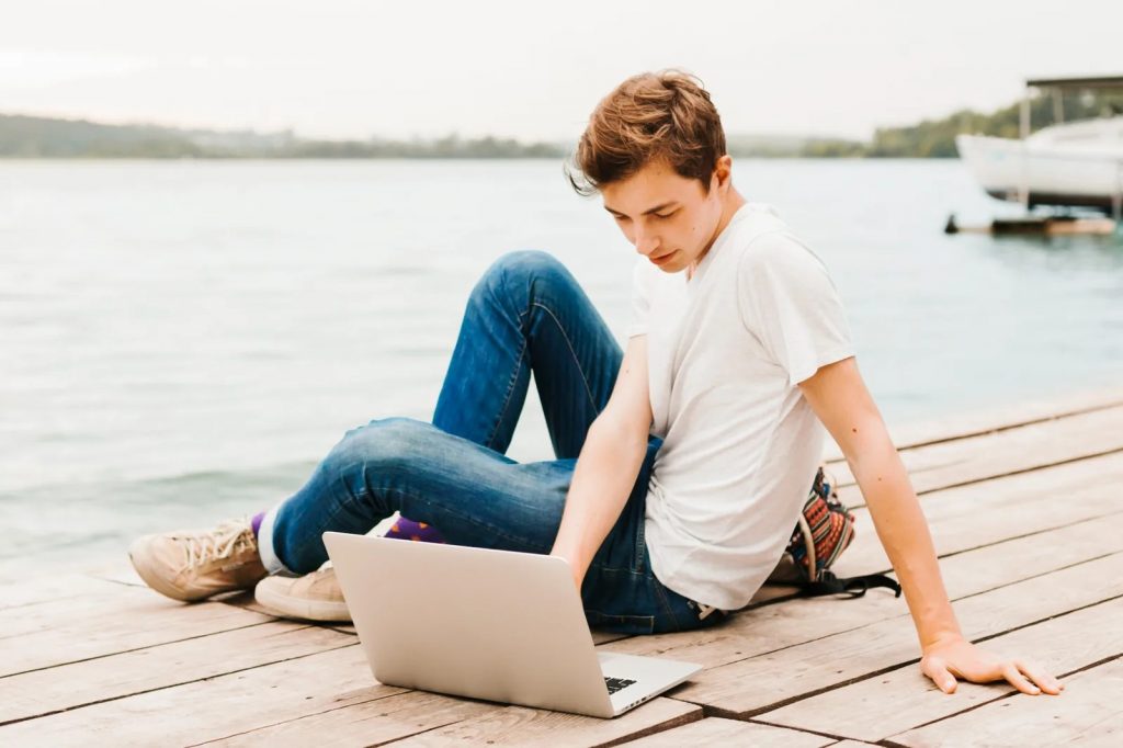 Un adolescent suit un cours particulier en ligne sur un ordinateur portable au bord d’un lac pendant l’été.